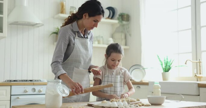 Happy pretty baker mother and sweet little daughter kid baking in home kitchen together, shaping, rolling dough, making pies, cookies, dumplings. Mother teaching girl in apron to cook bakery food