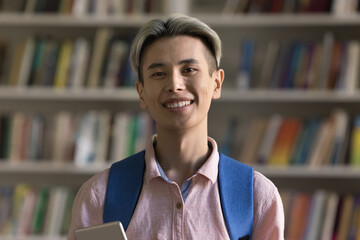 Head shot portrait Asian teenage guy with dyed blonde hair, wear backpack, holds tablet posing alone in university library smile look at camera. Excellent studies, get higher education, scholarship