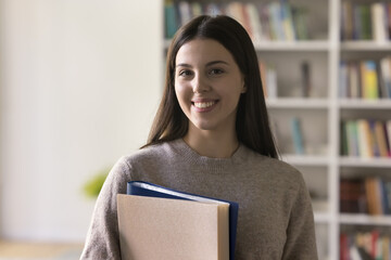 Head shot portrait of pretty teenager, student girl holding folders pose standing in campus library...