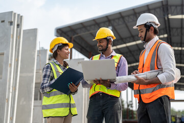 Team of construction engineers wearing vest and helmet safety discussing project at construction site. Group Indian foreman with laptop, paperwork working at factory making precast concrete wall.
