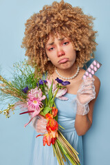 Upset curly haired woman with red eyes and nose has allergy to flowers holds bouquet and pills to cure disease wears festive dress and necklace poses against blue background. Allergic reaction