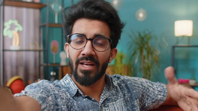 POV of indian man blogger taking selfie on smartphone, communicating video call online with social media subscribers followers, recording vlog stories. Young hindu guy at home apartment room on couch