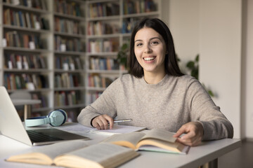 Portrait of pretty teen girl student studying in library, doing homework sit at desk smile staring at camera enjoy studies, gains new knowledge, practicing language, prepare for university admission