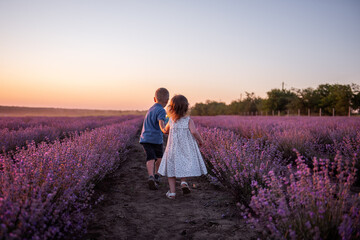Playful cute boy girl are playing in rows of lavender purple field at sunset. Small couple runs after each other, catches up, holding hands. Cheerful, happy childhood. Travel in countryside. Allergy