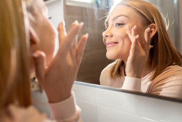 Smiling woman applies facial cream in her bathroom as part of her healthy morning routine.