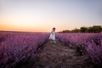 Obraz premium Little girl in flower dress runs across field of purple lavender among the rows at sunset. Toddler child have fun on walk in the countryside. Allergy concept. Natural products, perfumery. Copy space