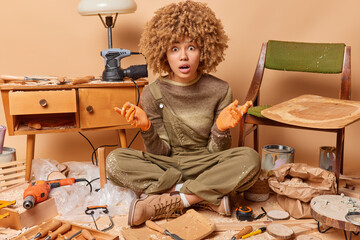 Fototapeta na wymiar Photo of stunned adult female carpenter being engaged in woodworking poses among joiner equipment sits in lotus pose produces wooden furniture wears jumper and overalls enjoys her profession