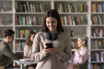 Teenage student girl standing in library or classroom use smart phone, search information, take break, read sms from friend, chatting on-line, check timetable through mobile apps. Modern tech, studies