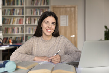 Beautiful smiling brunette teen girl posing in university library seated at desk with laptop and textbooks. Portrait of excellent student, higher education, successful, interesting high school studies