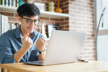 close up young asian man open laptop and sit in coffee shop while raise hand for celebration after winning reward on internet and lifestyle concept