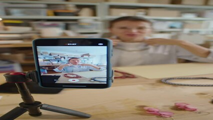 Vertical rack focus shot of woman showing handmade jewelry and telling about it on camera in workshop while filming video advertising or DIY vlog with smartphone on tripod
