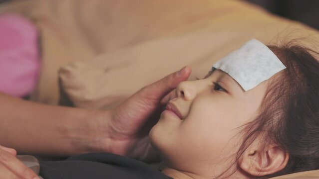 Close up face of adorable Asian girl who is sick and has fever, and lying down on her bedroom with her mother who is taking care with rubbing  towel to cooling down her temperature.