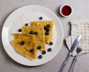 Three pancakes are on a white round plate, there are blueberries on the plate next to it. The pancakes are sprinkled with honey on top. Next to the plate there is a white bowl of honey, a white towel.