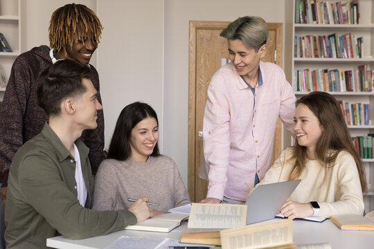 Five happy diverse teenage students gather together, smile, laugh, enjoy talk, distracted from studying, blab seated at desk stand in library, engaged in teamwork, share ideas and opinion over task - Powered by Adobe
