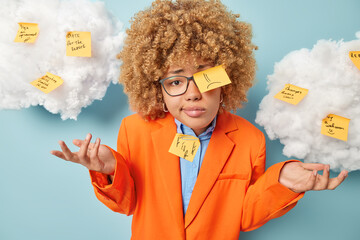 Hesitant clueless businesswoman covered with sticky notes shrugs shoulders spreads palms writes down list to do dressed in orange jacket isolated over blue background with white clouds overhead