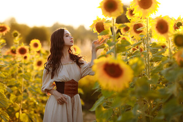 A beautiful young tall slender brunette woman in a beige dress and leather corset stands in a field with blooming yellow sunflowers at sunset. 