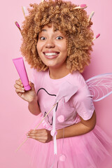 Vertical shot of cheerful curly haired female tooth fairy holds toothpaste to clean teeth and wand...