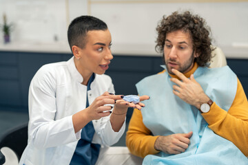 Young woman professional dentist showing the details of dental impression to a male patient, preparing him for dental restorations, explaining upcoming dental work for healthy gums and teeth.