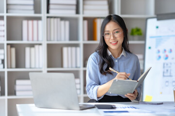 Happy young Asian businesswoman sitting at desk and take notes with laptop computer in the office.