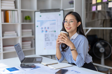 Happy young asian businesswoman sitting working at office and drinking coffee.