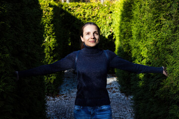 Portrait beautiful young woman in blue sweater posing against backdrop of green hedge. Woman with arms spread apart walking in public park at sunny day