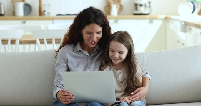 Happy engaged mom and cute little daughter kid watching cartoon movie on laptop, hugging on couch, using computer for online internet communication, entertainment, enjoying leisure