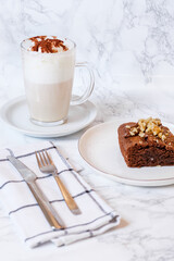 Latte macchiato specialty coffee with cocoa powder, mocha and foam over white marble  background and plate served with a chocolate brownie with nuts, spoon and fork.