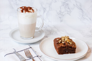 Latte macchiato specialty coffee with cocoa powder, mocha and foam over white marble  background and plate served with a chocolate brownie with nuts, spoon and fork.