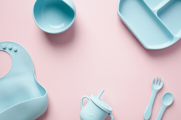 Flat lay silicone bib, plate, cup, cutlery on pastel pink background. Serving baby food concept.