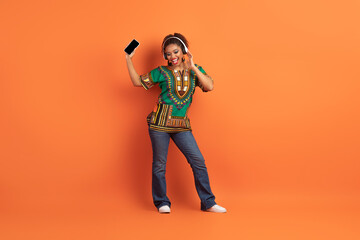 Fototapeta na wymiar Joyful black woman in african clothes using headphones and smartphone