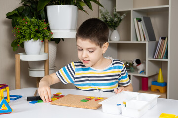 A preschool child plays with a mosaic, builds figures on a board, hammering nails with a hammer on multi-colored parts of the designer