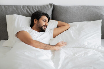 Young middle eastern guy in white t-shirt sleeps, lies on bed with empty space in bedroom interior, top view