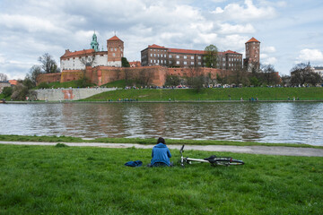 Lonely relaxing woman with bike is sitting on grass by Vistula River opposite to the Wawel Royal...