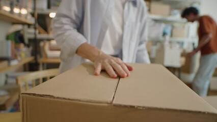 Cropped shot of woman sealing cardboard box with transparent adhesive tape while preparing package for shipping order in delivery service office