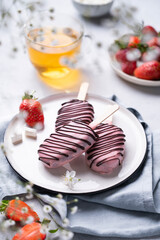 Chocolate cake pops ice cream on a stick in pink glaze on a plate with berries on a light background with cup of tea. Healthy homemade sweets.