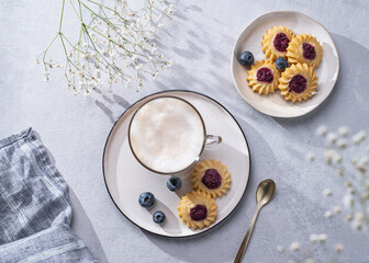 Cappuccino or latte with milk foam in a cup with handmade berry cookies and blueberries on a light background with gypsophila branches and shadow.