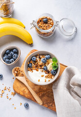 Homemade baked granola with yogurt, blueberries and banana in a bowl on a light background with fresh berries. Healthy vegetarian breakfast with muesli.