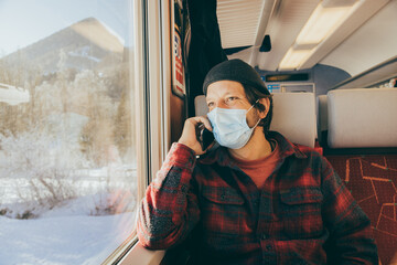 Fototapeta na wymiar Portrait of caucasian young man wearing a protective face mask on train travelling sitting next to the window and talking on the phone.