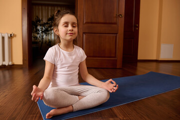 Cute conscious 5-6 years old child girl, practicing yoga indoors, sitting on a blue fitness mat with her eyes closed, meditating in lotus pose and mudra gesture. Mindfulness. Meditation. Yoga practive - obrazy, fototapety, plakaty