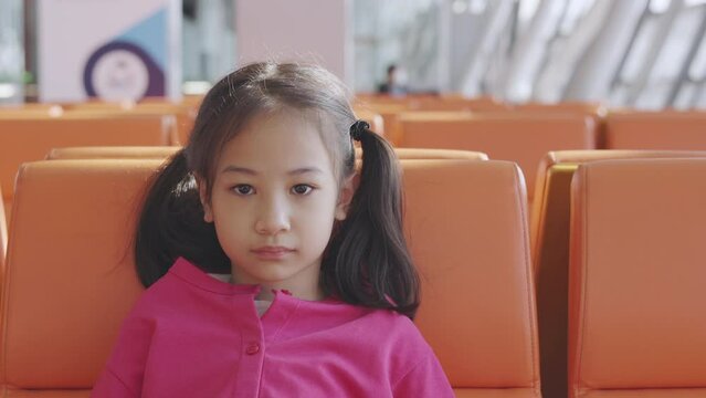 Close up face of adorable asian kid girl who is sitting on bench at airport terminal alone shows concept of waiting, lost, unhappy or depressed emotion of child with beautiful sunlight through window.