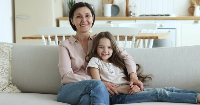 Happy loving grandma and granddaughter kid looking at camera, resting on sofa. Grandmother hugging grandkid in home living room, laughing, smiling, enjoying family leisure time