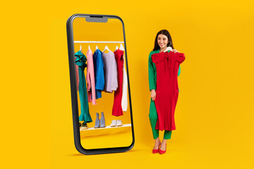Excited shopaholic lady holding red dress and looking at cellphone display with clothing rail,...