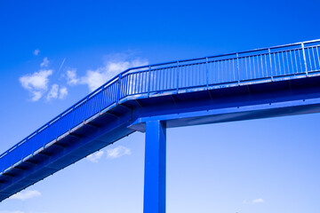 A modern pedestrian overpass across a highway. Blue bridge without people against blue sky in summer day. Minimalistic architecture, urban infrastructure. Bridge on stilts high above a ground.