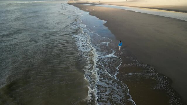 Young girl on sandy coast. Girl leaves footprints on sand. Attractive woman in blue sweater on beach. Girl walking along seashore. Aerial view of seashore. Beautiful ocean waves. Beach with  sand