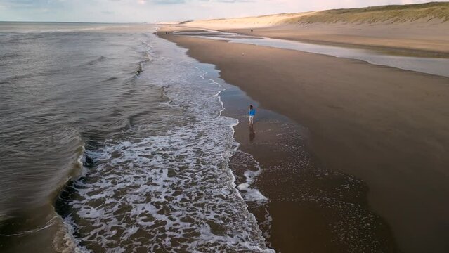Young girl on sandy coast. Girl leaves footprints on sand. Attractive woman in blue sweater on beach. Girl walking along seashore. Aerial view of seashore. Beautiful ocean waves. Beach with  sand