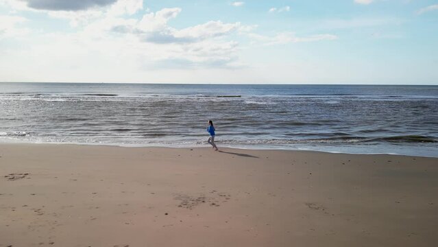 Young girl on sandy coast. Girl leaves footprints on sand. Attractive woman in blue sweater on beach. Girl walking along seashore. Aerial view of seashore. Beautiful ocean waves. Beach with  sand