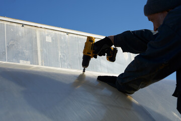 A man attaches a polycarbonate sheet to the metal frame of a greenhouse. He tightens a self-tapping...