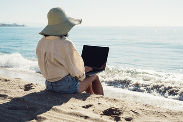 young beautiful girl freelancer with a laptop works on the seashore sitting on the sand