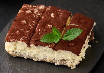 A piece of tiramisu sprinkled with cocoa, on top of a sprig of fresh mint on a black background