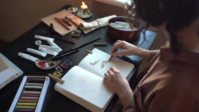 Woman paintress draws picture using pastel chalks sits at table in art studio. Girl painting in sketchbook practicing paint skills. Romantic atmosphere with burning candle. University art student.